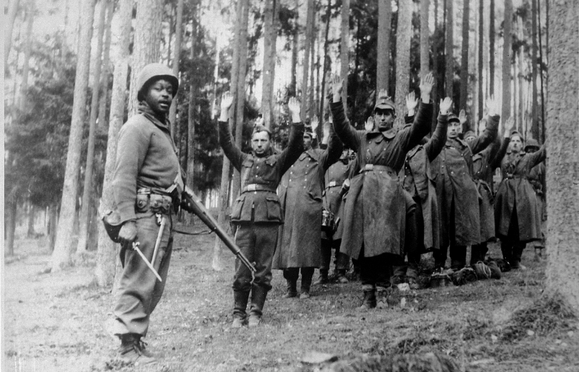 A Black soldier of the 12th Armored Division stands guard over a group of Nazi prisoners, April 1945.